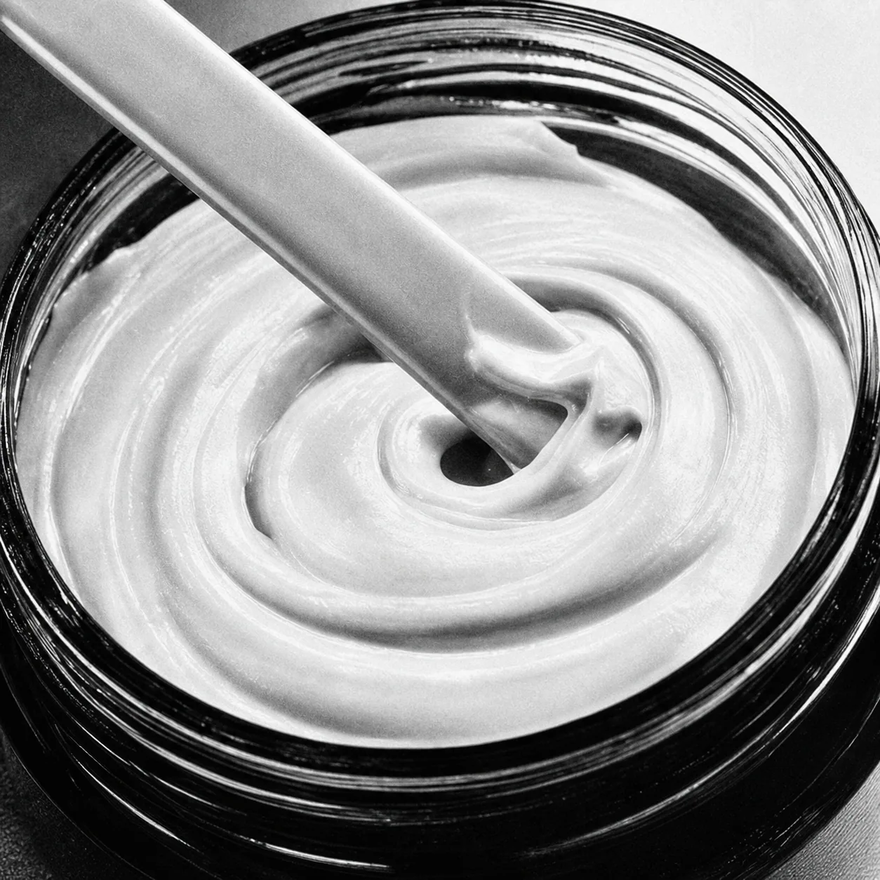 A spatula stirring a cream formula in a glass jar